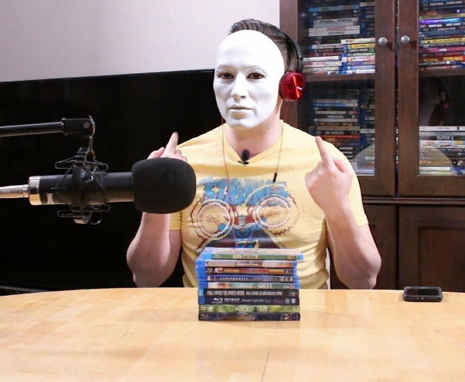 Tim Kuefler wearing a white mask and headphones, sitting at a podcasting table — referencing his mascot origin story